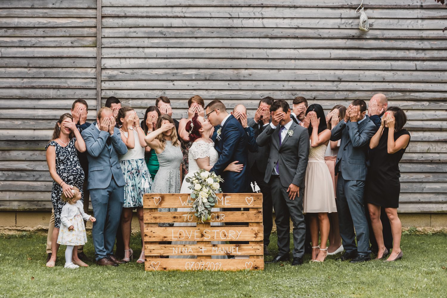 Gruppenfoto Hochzeitsgesellschaft bei der sich Brautpaar küsst und alle anderen die Augen zu halten.